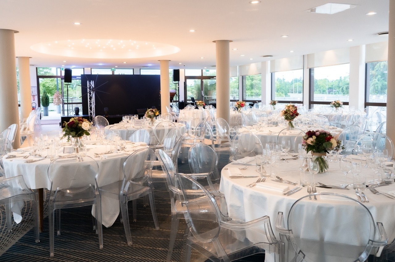 Salon Plaisance Salle de réception lumineuse avec tables rondes dressées, chaises transparentes et bouquets de fleurs au centre.