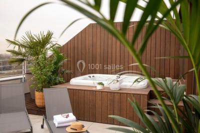Jacuzzi extérieur entouré de plantes et de chaises longues sur une terrasse avec un panneau en bois.
