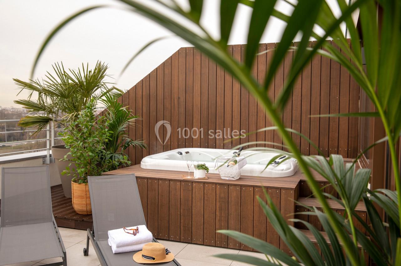Jacuzzi extérieur entouré de plantes et de chaises longues sur une terrasse avec un panneau en bois.