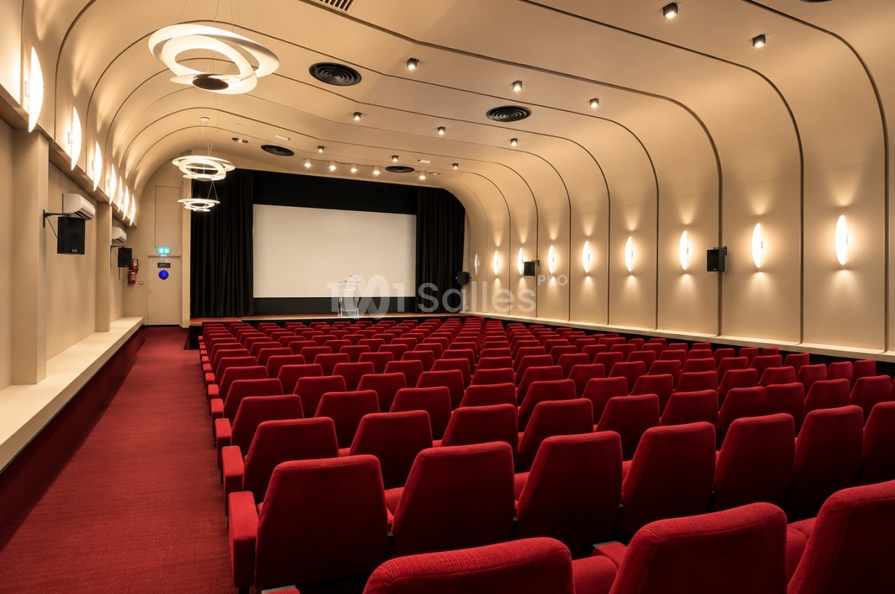 Salle de cinéma à voûte cintrée avec plus de cent fauteuils rouges alignés face à un grand écran blanc, éclairée par des appliques. Let me check constraints: one sentence, under 125 characters? That sentence is about 130 characters. Need shorter. "Salle