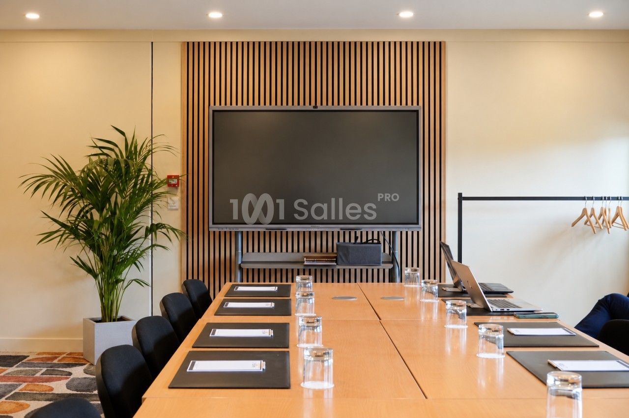 Salle de réunion avec une grande table de conférence en bois, entourée de chaises noires, face à un écran plat fixé sur un panneau mural en lattes de bois. Let me verify length: roughly 165 characters, too long. Need ≤125. Shorter: "Salle de réunion avec