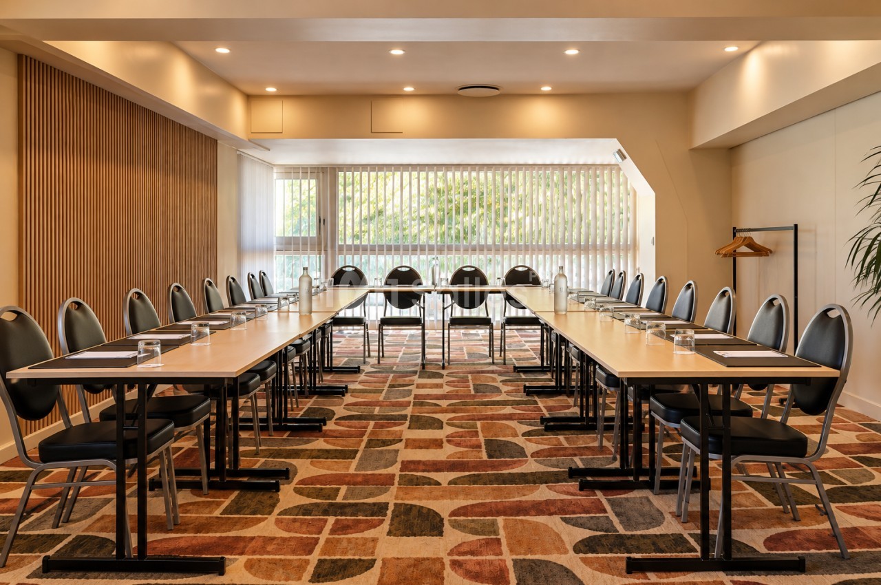 Salle de réunion en configuration en U avec tables blanches, chaises noires à dossier arrondi, carafes et verres, éclairée par des spots au plafond et une grande baie vitrée. Let me check length: roughly 165 characters, too long. Need ≤125. Salle de