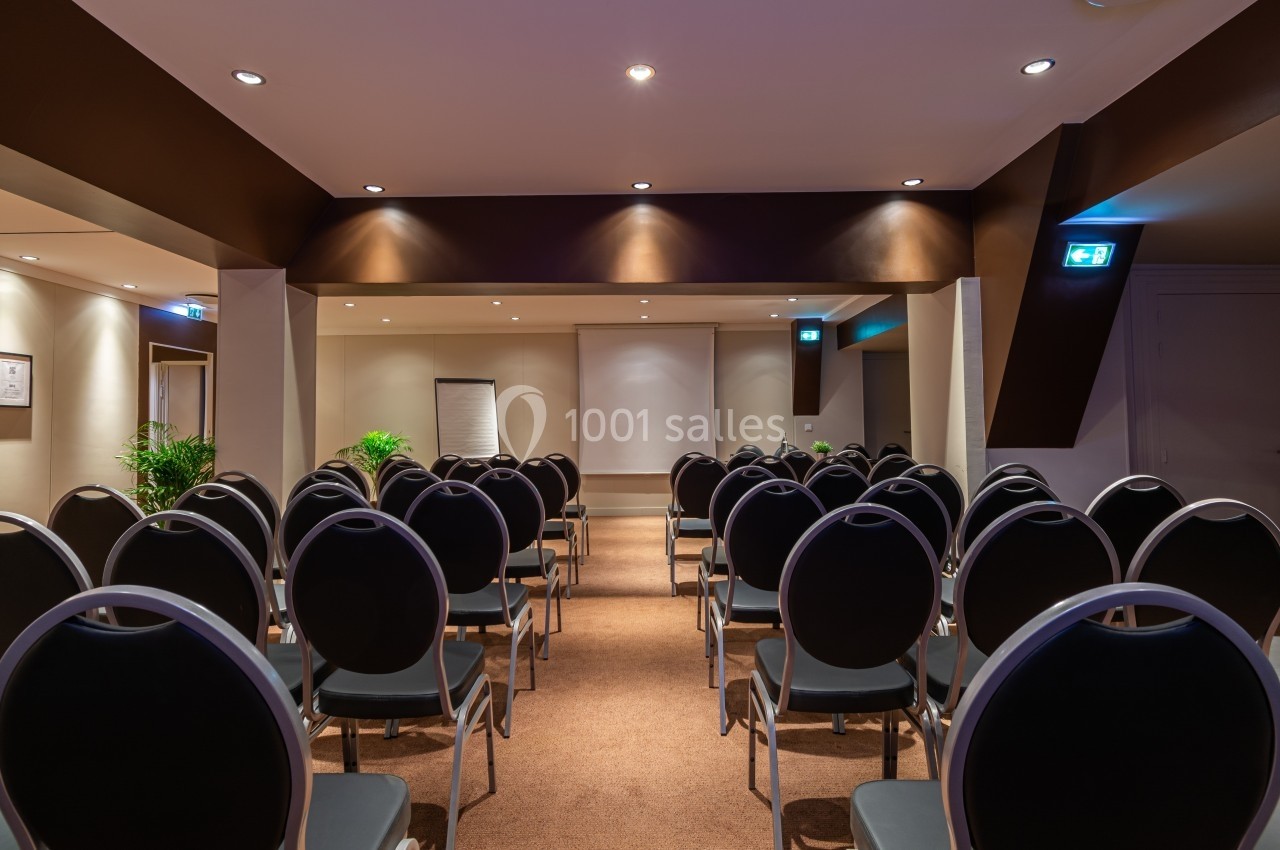 Salle de conférence avec des rangées de chaises noires, un écran blanc, un tableau de présentation et des plantes…