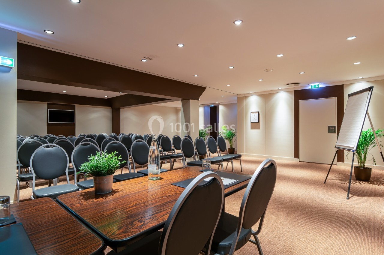 Salle de réunion avec chaises alignées, tables en bois, plantes décoratives et un tableau de présentation.