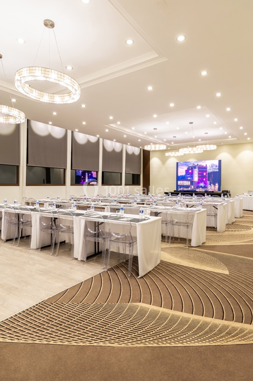 Salle de conférence lumineuse avec des tables alignées, des chaises transparentes et un écran de projection allumé.