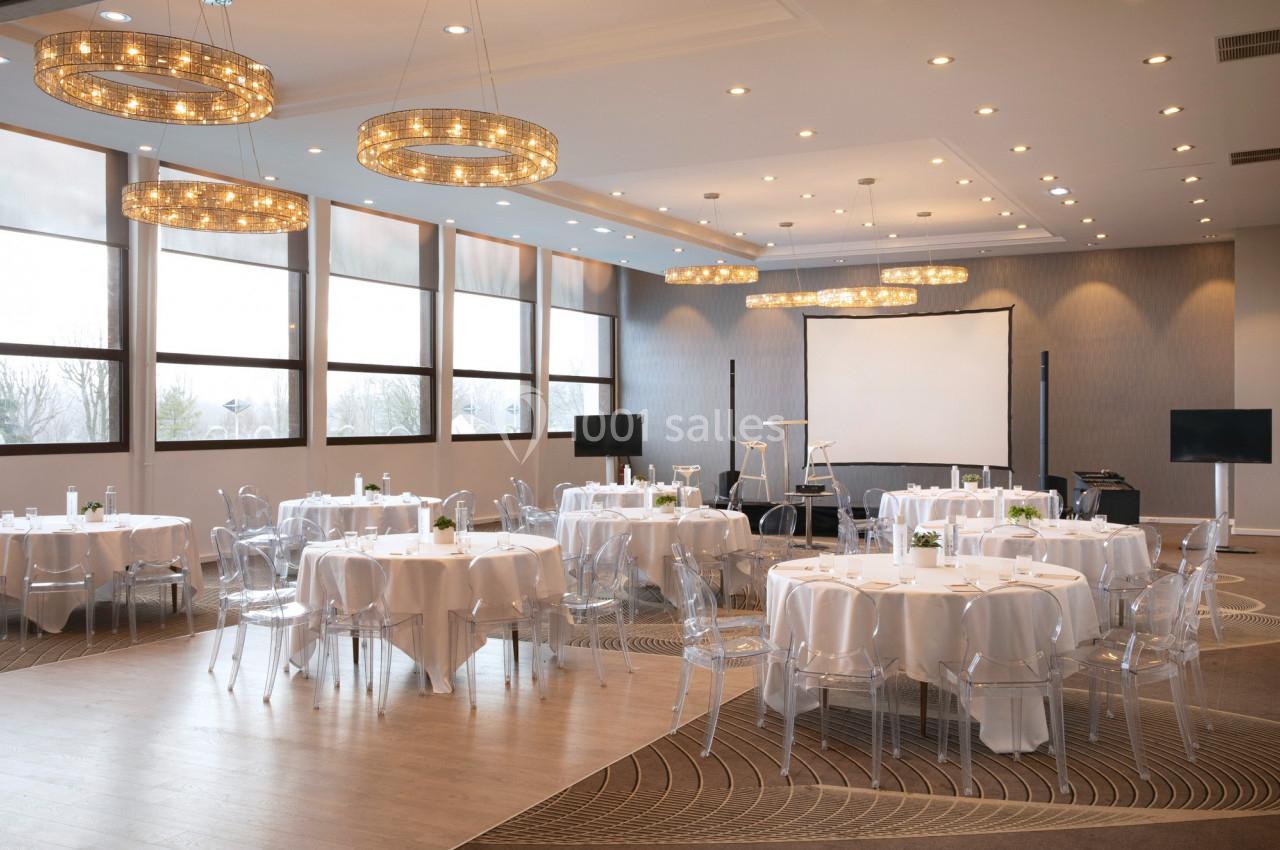 Salle de réception lumineuse avec tables rondes dressées, chaises transparentes et écran de projection en fond.
