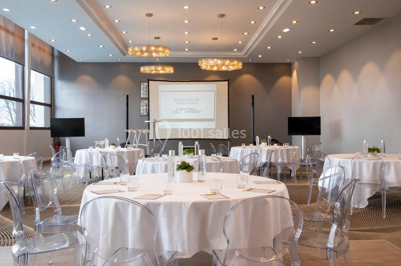 Salle de conférence lumineuse avec tables rondes dressées, chaises transparentes et écran de projection allumé.