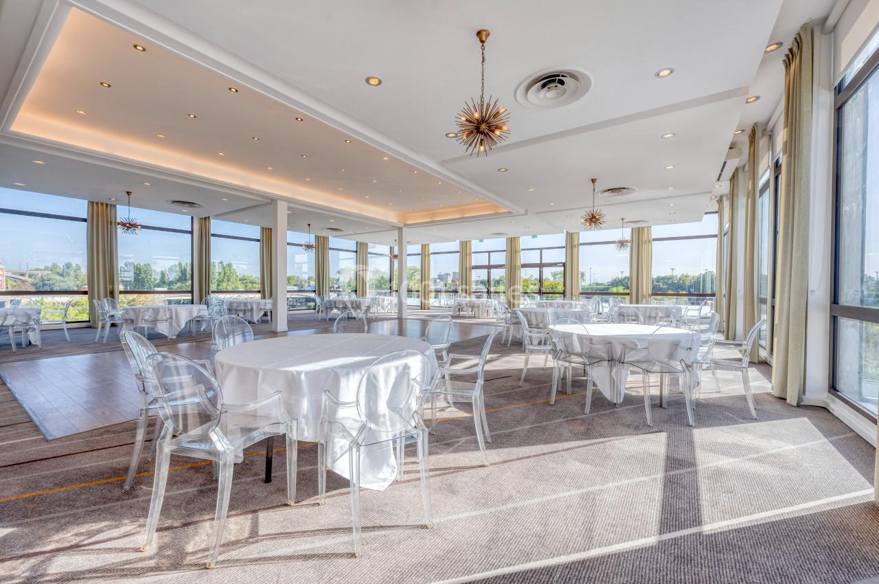 Salle lumineuse avec de grandes baies vitrées, tables rondes dressées de nappes blanches et chaises transparentes.