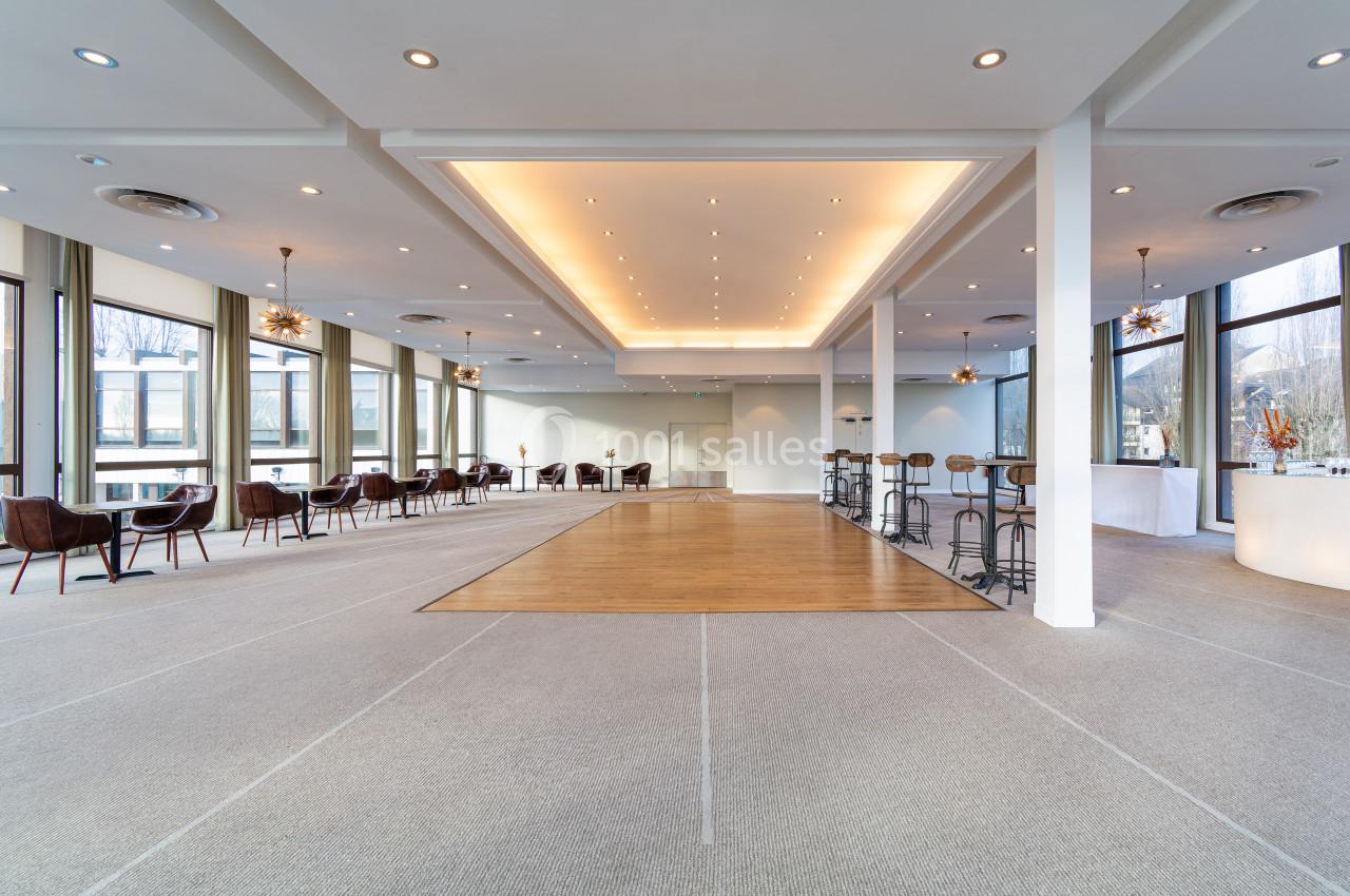 Salle lumineuse avec de grandes baies vitrées, un sol en moquette et une zone centrale en parquet, aménagée avec des tables…