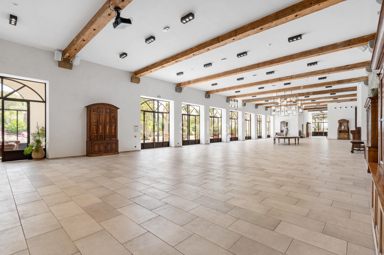 Grande salle lumineuse avec sol carrelé, poutres apparentes au plafond et grandes fenêtres donnant sur l'extérieur.