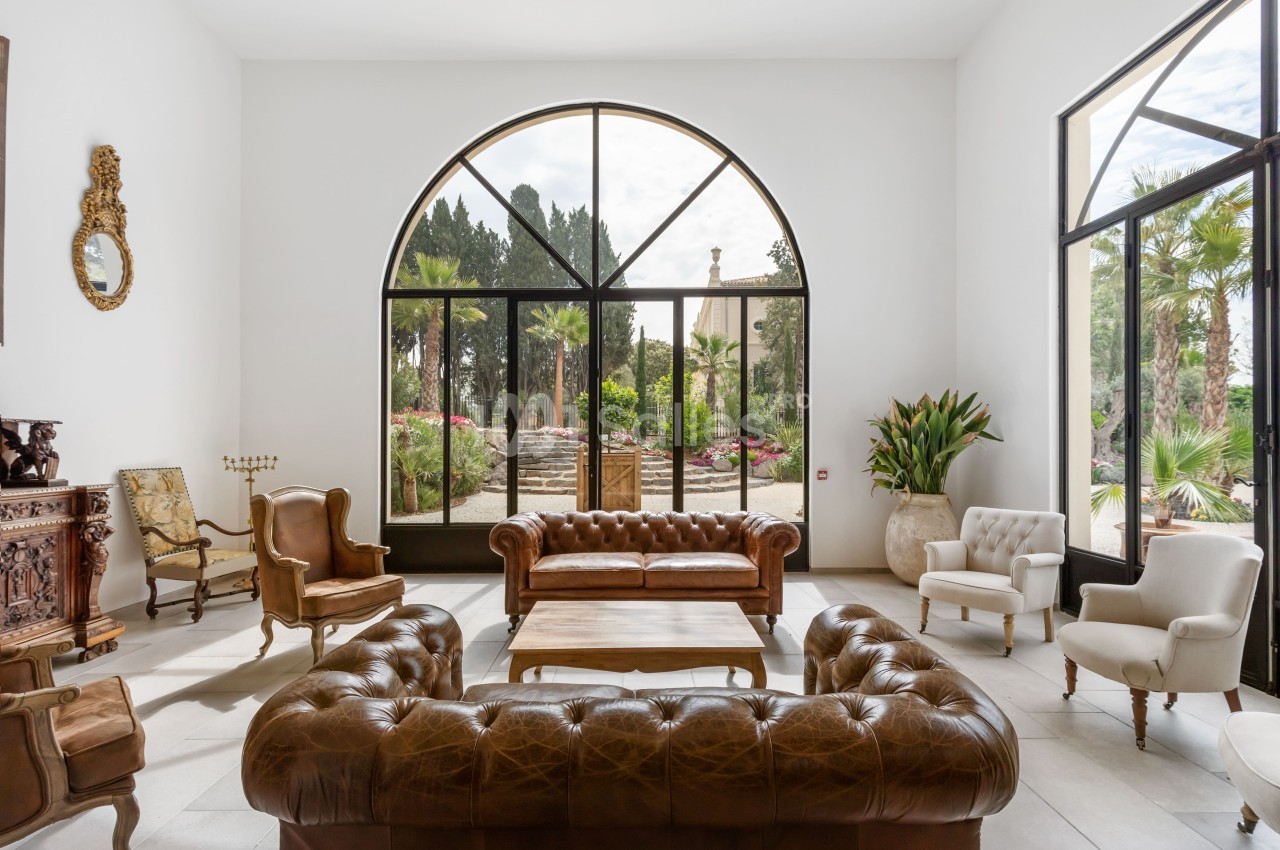 Salon lumineux avec canapés en cuir, fauteuils, table basse en bois et grandes baies vitrées donnant sur un jardin.