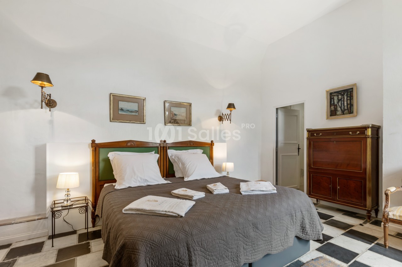 Chambre avec un grand lit, linge de lit plié, mobilier ancien, murs blancs et sol en carreaux noir et blanc.