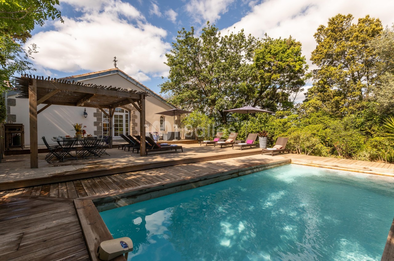 Piscine extérieure entourée de transats et d'une terrasse en bois, avec une pergola et une maison en arrière-plan.