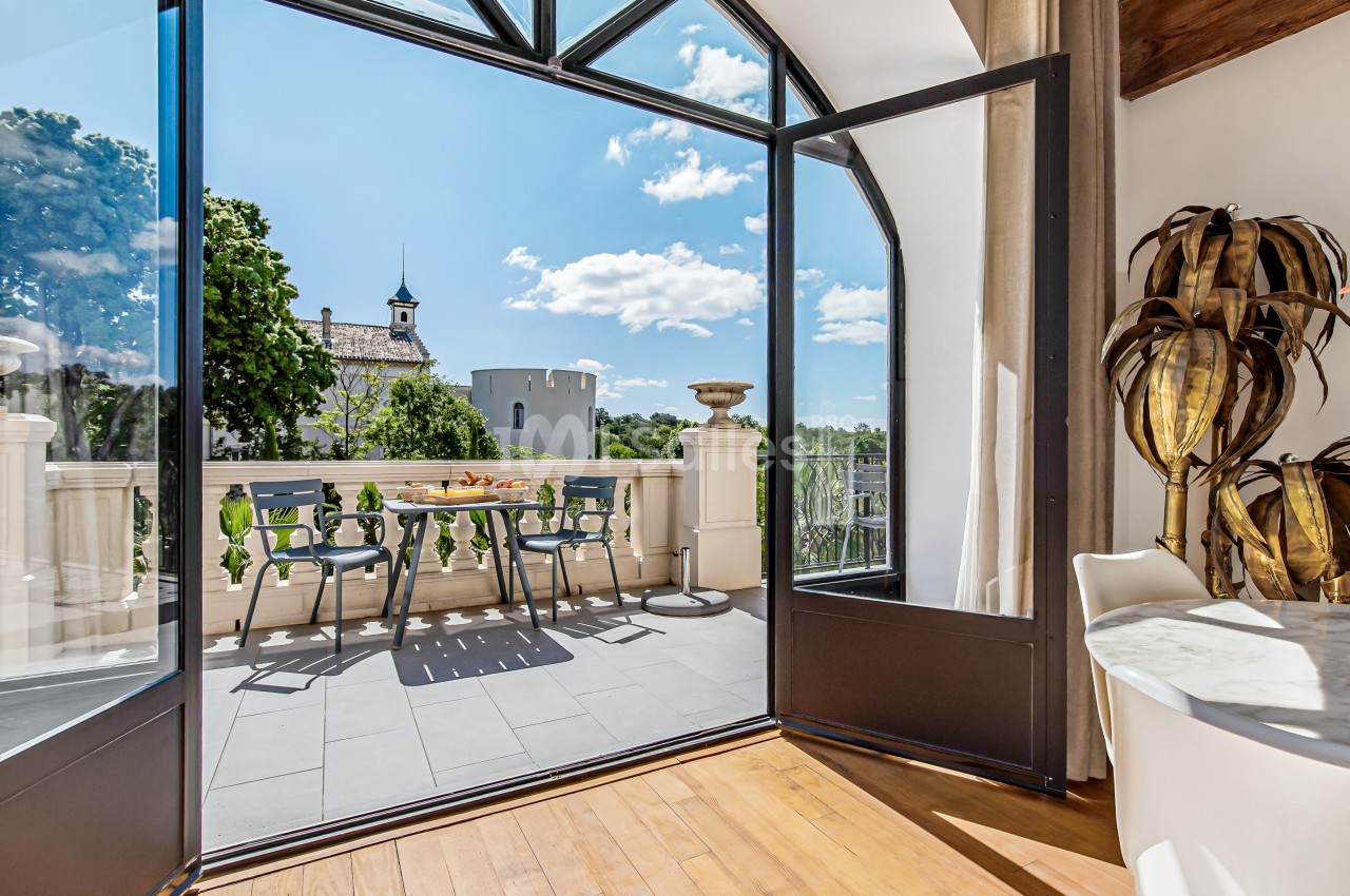 Vue depuis une pièce lumineuse sur une terrasse aménagée, avec table et chaises, donnant sur un paysage arboré et une tour.
