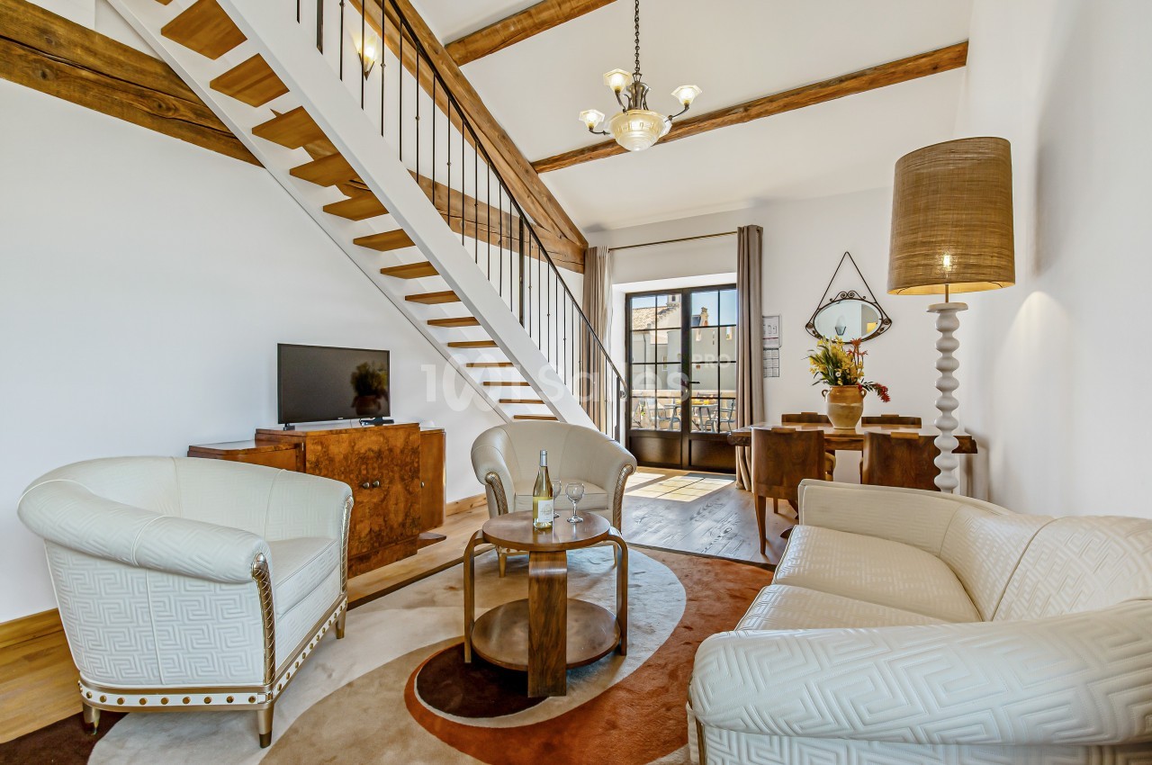 Salon lumineux avec mobilier classique, escalier en bois, plafond à poutres apparentes et grande fenêtre donnant sur l…