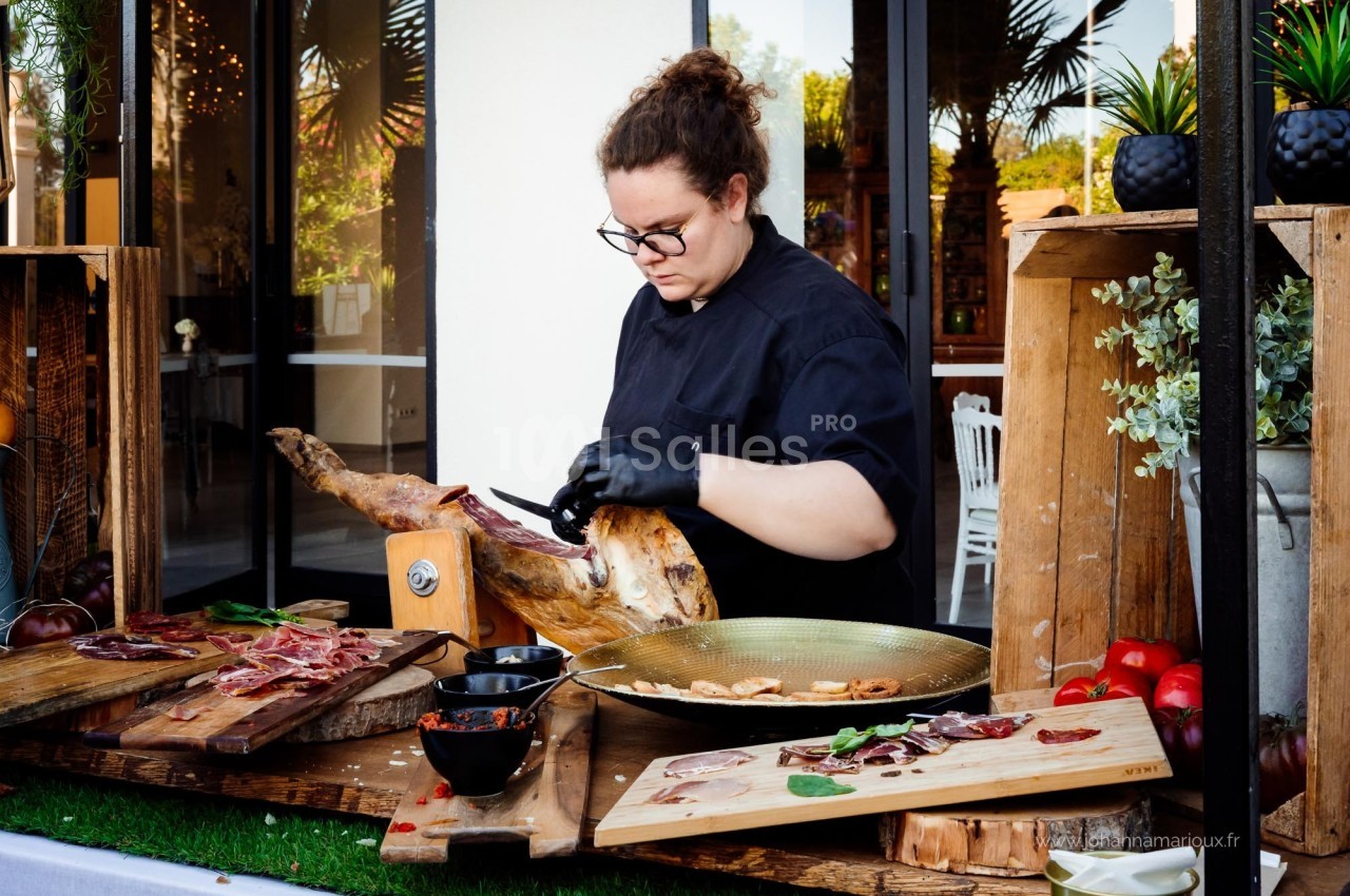 Une personne découpe un jambon sur un support en bois, entourée de planches garnies de charcuterie et de légumes.