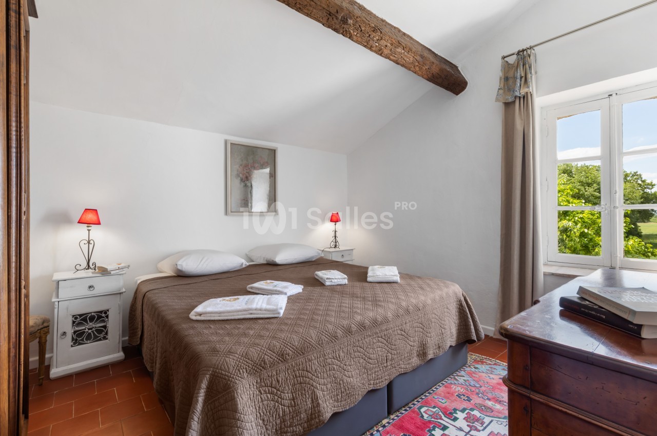 Chambre lumineuse avec lit double, couvre-lit marron, poutre apparente, fenêtres ouvertes sur un jardin verdoyant.