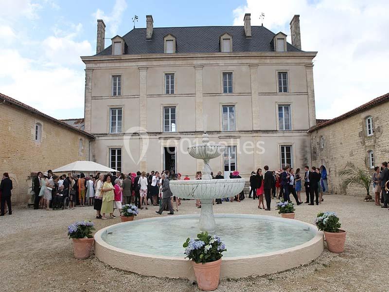 Façade d'un château en pierre avec une fontaine au premier plan et des invités rassemblés dans la cour.