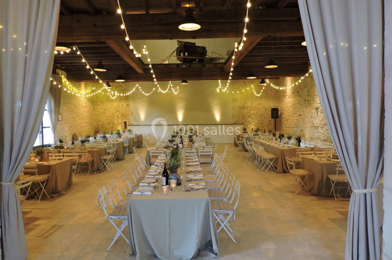 Salle de réception décorée avec des guirlandes lumineuses, des tables dressées et des chaises blanches pliantes.