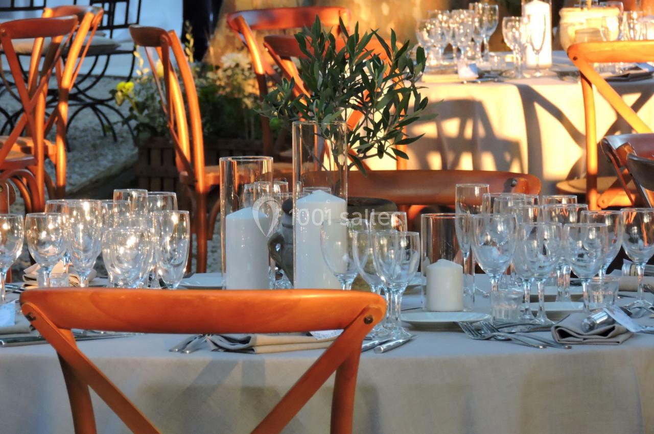 Table dressée en extérieur avec nappes blanches, chaises en bois, verres, bougies et décoration végétale.