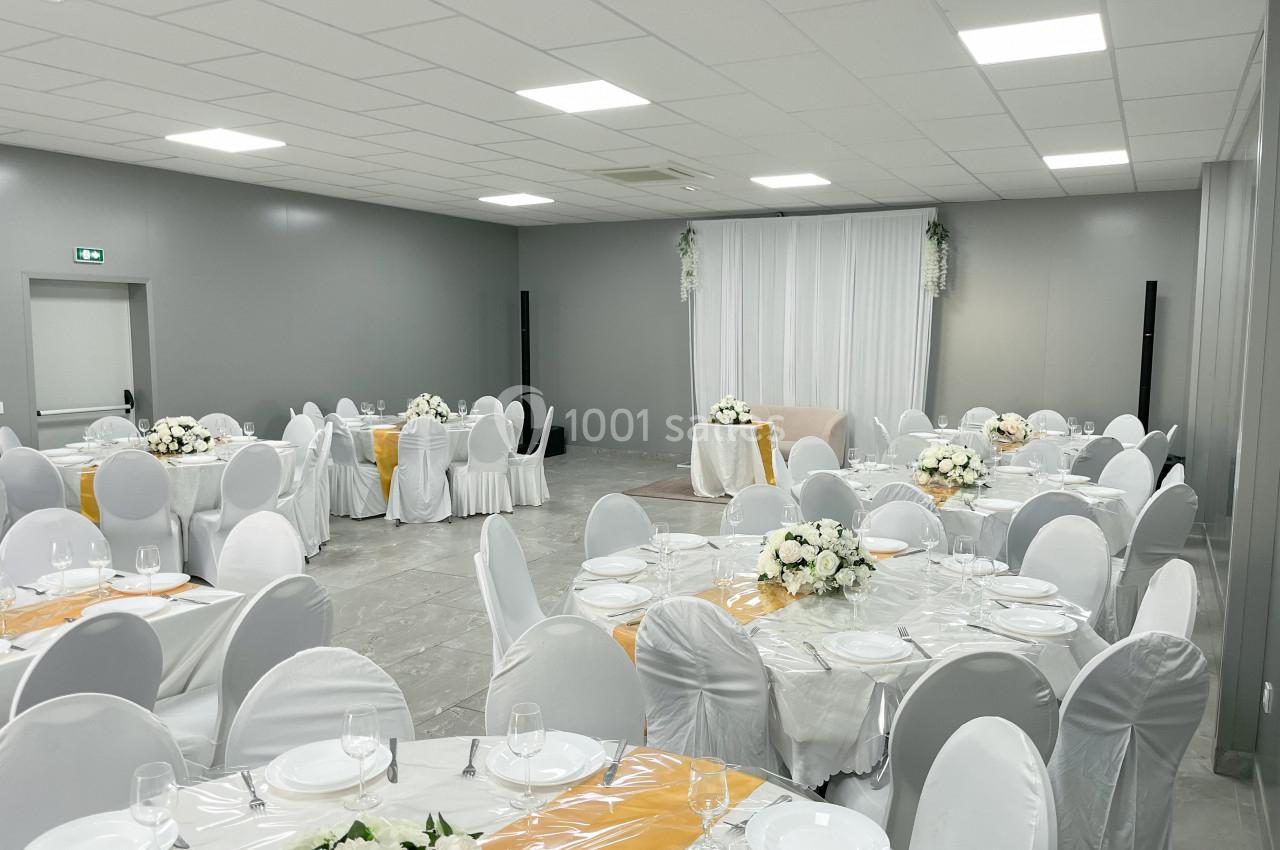 Salle de réception décorée avec des tables rondes, nappes blanches, chaises houssées et compositions florales.