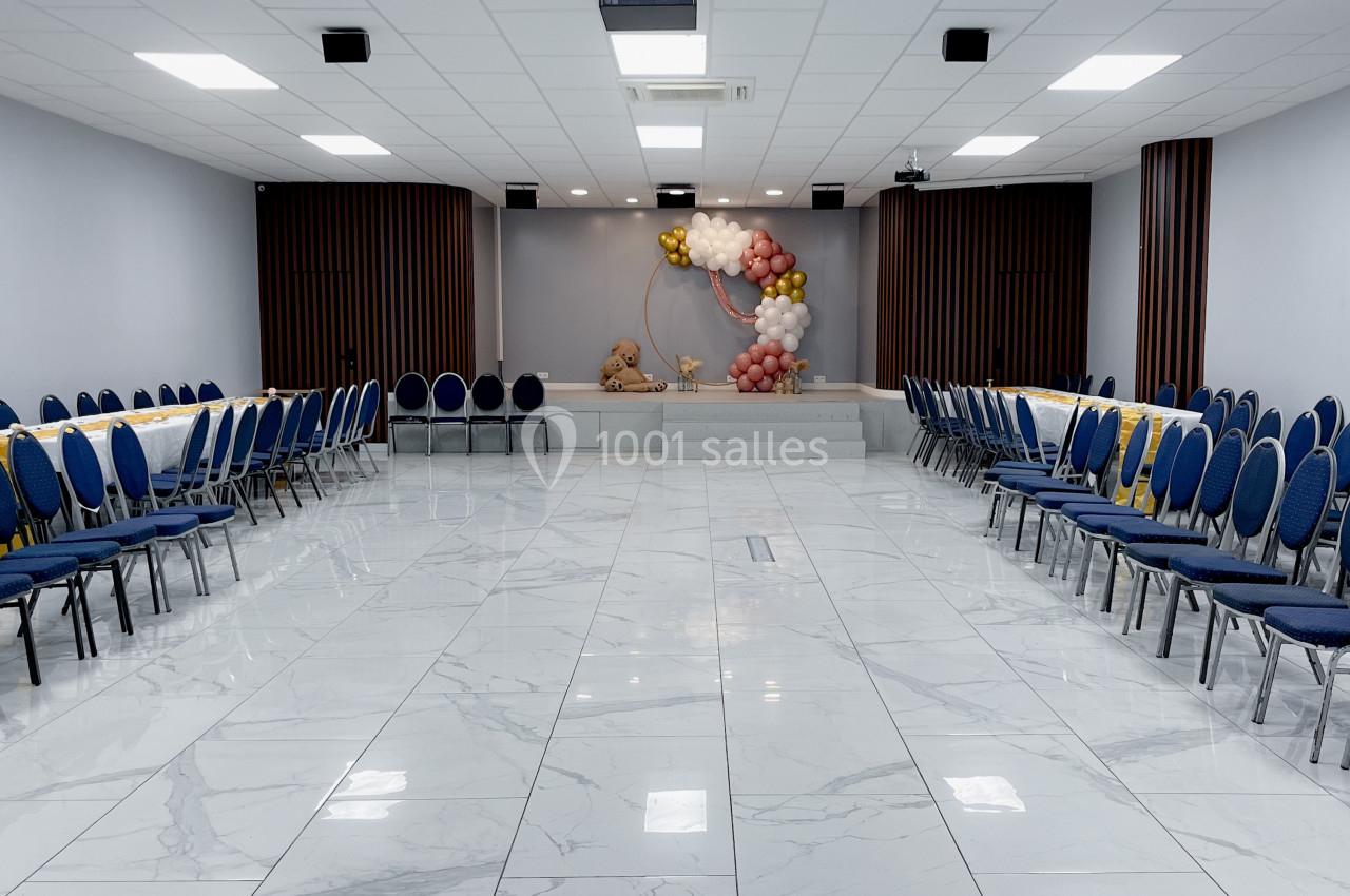 Salle de réception lumineuse avec sol en marbre, chaises bleues alignées et décoration florale sur une estrade.