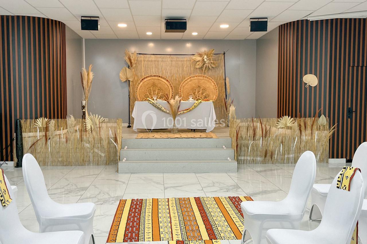 Salle décorée pour un événement avec des chaises blanches, tapis colorés et scène ornée d'éléments naturels.