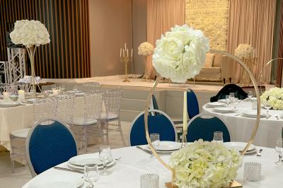 Salle de réception avec tables rondes couvertes de nappes blanches, chaises bleues et une scène avec rideaux bleus.