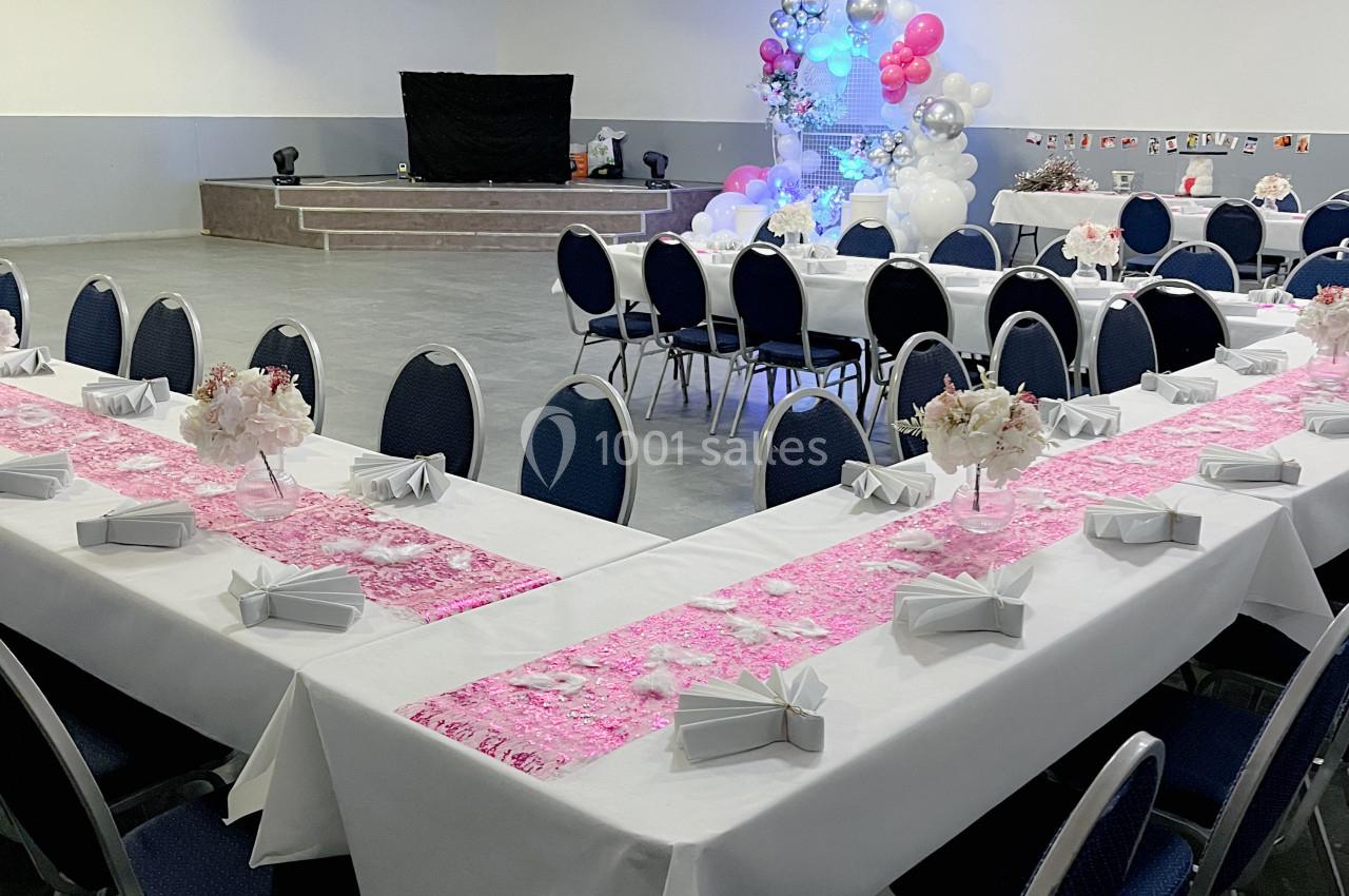 Salle de réception décorée avec des tables en U, nappes blanches, chemins de table roses et ballons en arrière-plan.