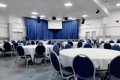 Salle de réception avec tables rondes couvertes de nappes blanches, chaises bleues et une scène avec rideaux bleus.