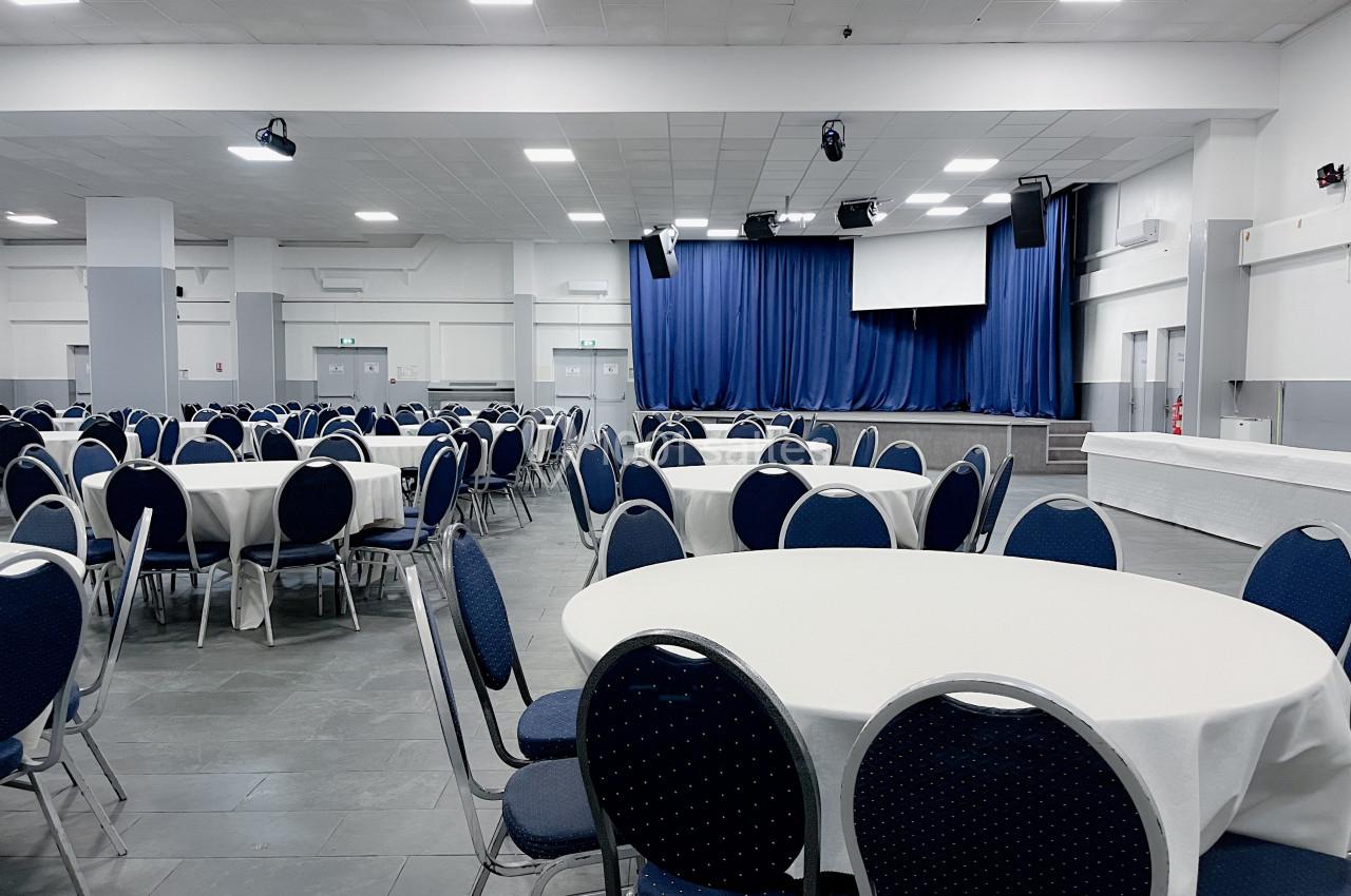Salle de réception avec tables rondes couvertes de nappes blanches, chaises bleues et une scène avec rideaux bleus.