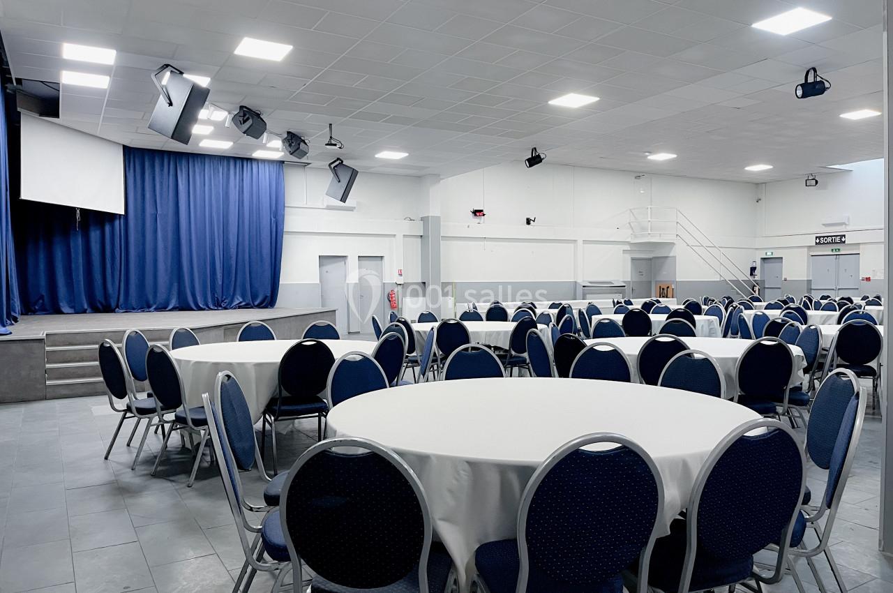 Salle de réception avec des tables rondes dressées de nappes blanches, une scène avec rideaux bleus en arrière-plan.