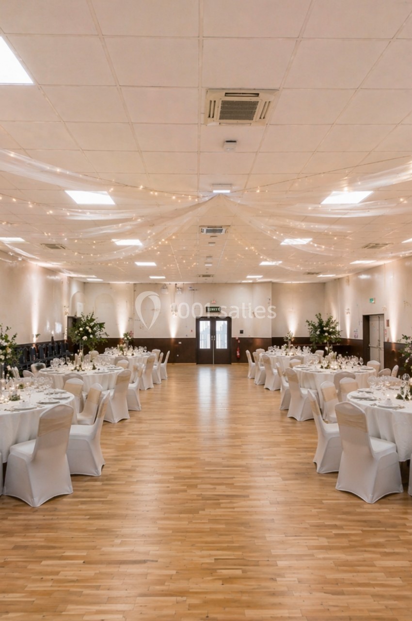 Salle de réception décorée avec des tables rondes dressées, chaises blanches et guirlandes lumineuses suspendues.