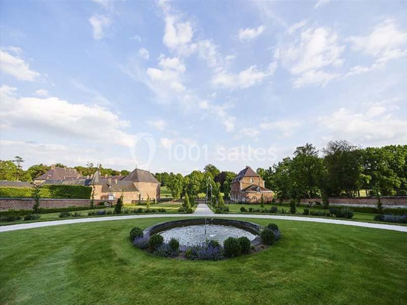 Vue d'un jardin paysager avec pelouse, fontaine centrale, allées circulaires et bâtiments en arrière-plan.