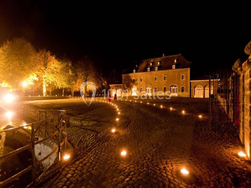 Cour pavée éclairée par des bougies et lumières chaudes, avec un bâtiment historique en arrière-plan, de nuit.