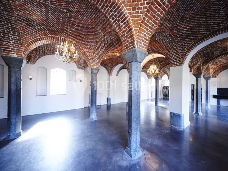 Salle voûtée en briques avec colonnes en pierre, sol en béton lisse et lustres suspendus.