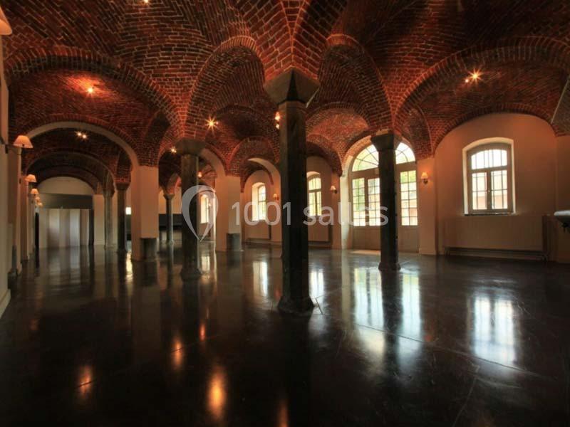 Salle voûtée en briques avec colonnes, sol sombre et grandes fenêtres laissant entrer la lumière naturelle.