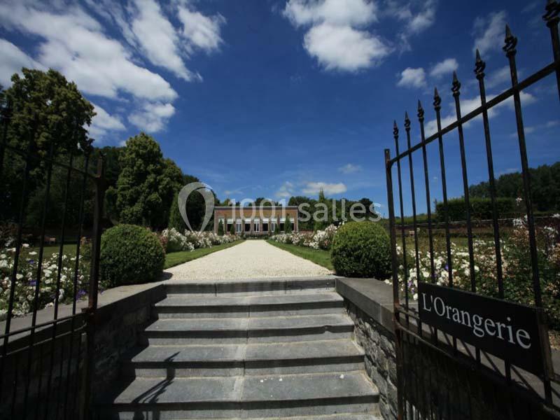 Entrée d'un jardin avec des grilles ouvertes, un chemin bordé de fleurs menant à un bâtiment nommé ’L'Orangerie’.