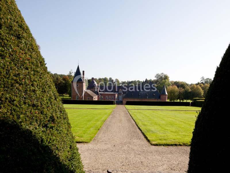 Allée centrale bordée de haies menant à un château en briques entouré de pelouses et d'arbres.