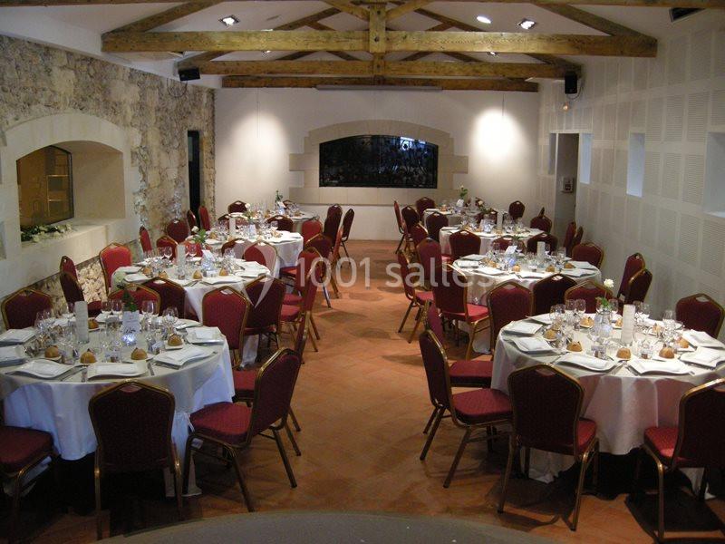Salle de réception avec tables rondes dressées, nappes blanches, chaises rouges et poutres apparentes au plafond.