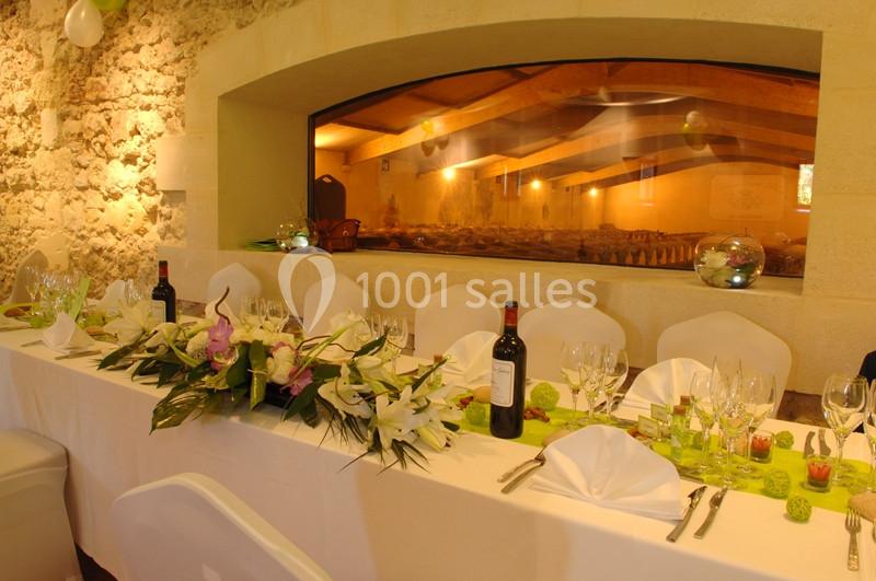 Table de réception élégamment dressée avec nappes blanches, fleurs, bouteilles de vin et vue sur une salle voûtée en arrière…