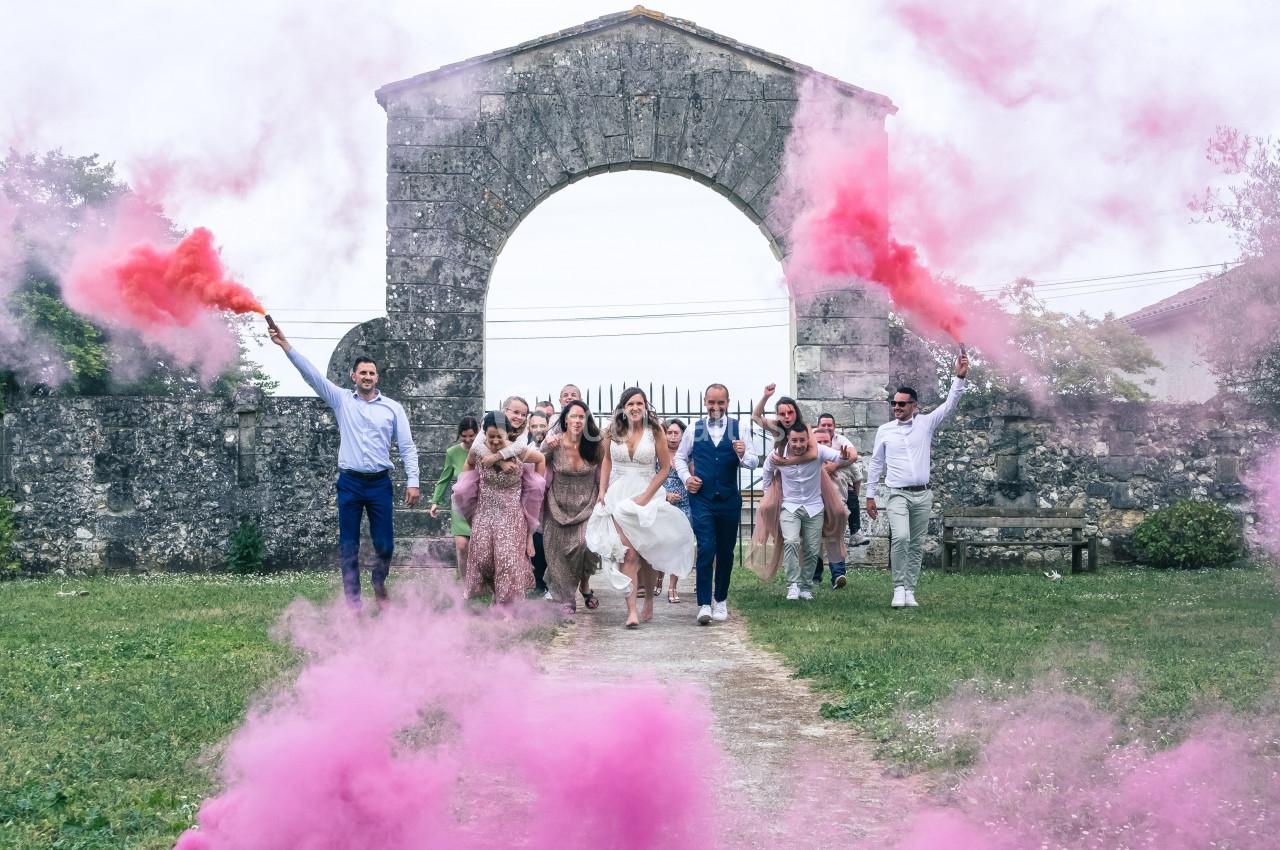 Un groupe de personnes en tenue festive court sous une arche en pierre, entouré de fumée rose.