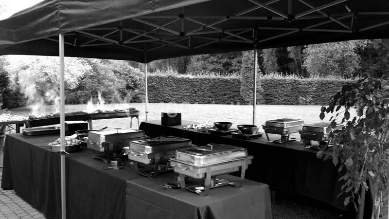 Buffet extérieur sous une tente avec des plats chauds en inox, installé dans un jardin avec haies en arrière-plan.