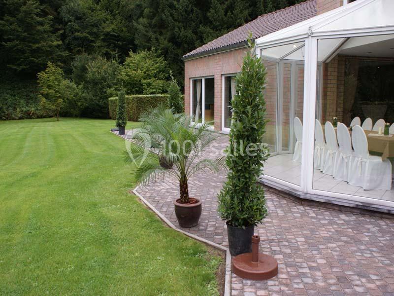 Terrasse pavée avec plantes en pot, bordant une maison en briques et une véranda, entourée de pelouse et d'arbres.