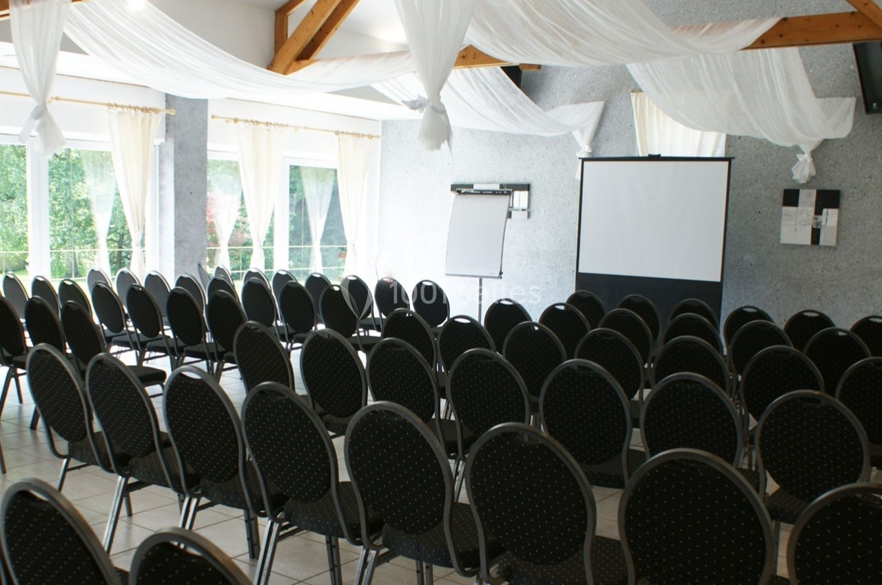 Salle de conférence lumineuse avec des rangées de chaises noires, un écran de projection et un tableau blanc.