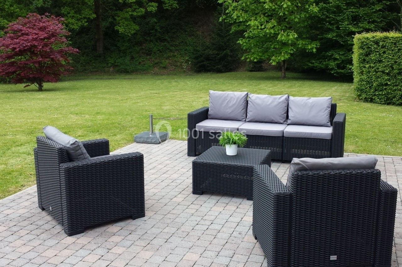 Mobilier de jardin en résine tressée avec coussins gris, disposé sur une terrasse pavée entourée de verdure.