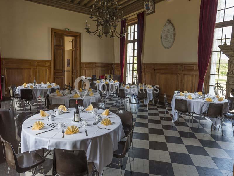 Salle de réception avec tables rondes dressées, nappes blanches, serviettes pliées jaunes et lumière naturelle.