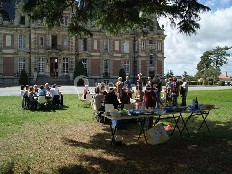Groupe de personnes partageant un repas en plein air devant un bâtiment ancien sur une pelouse ombragée.