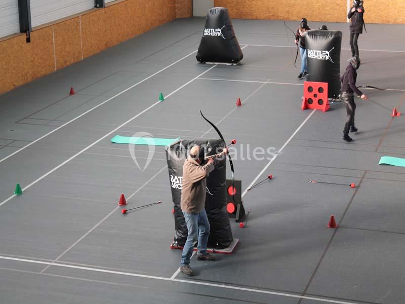 Un homme tire à l'arc dans une salle de sport aménagée avec des obstacles gonflables et des cibles.