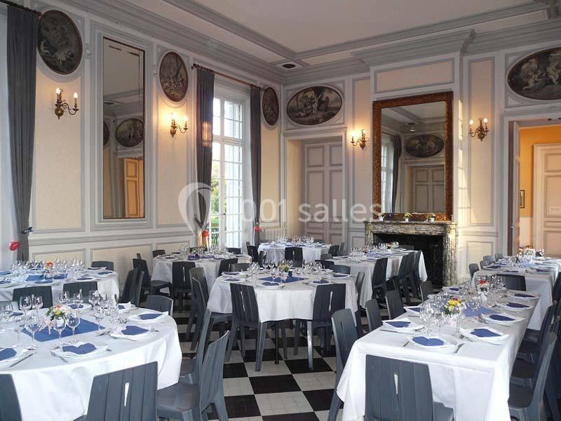 Salle élégante avec tables rondes dressées, nappes blanches, serviettes bleues, miroirs et moulures décoratives.